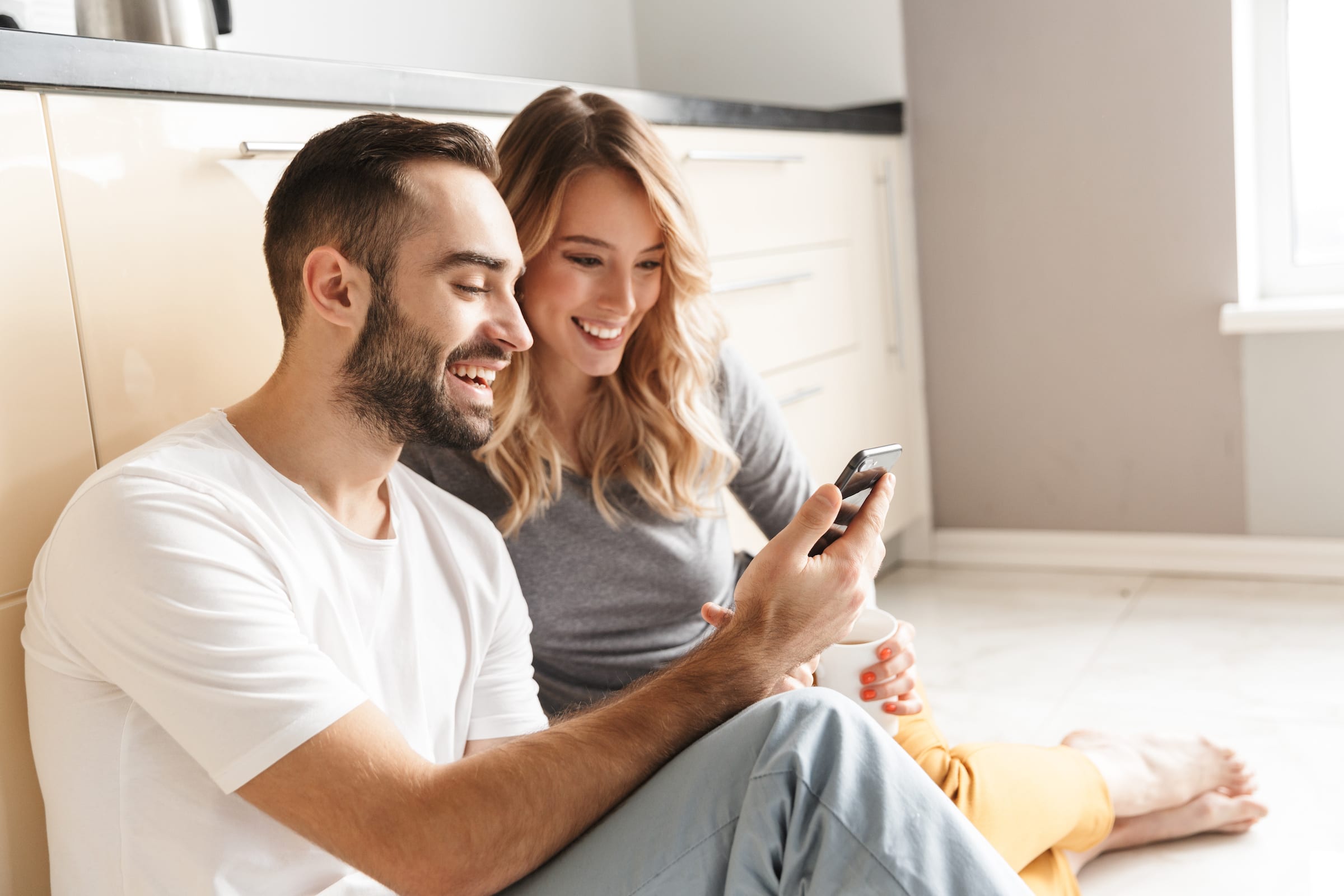 Image of amazing young loving couple sitting at the kitchen using mobile phone on a floor.